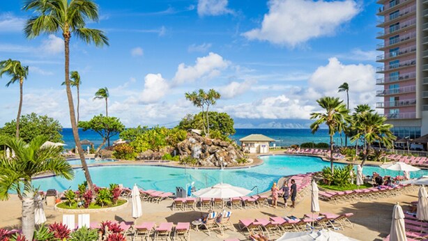 Ein Poolbereich eines Resorts mit Palmen, Liegestühlen und Blick aufs Meer.
