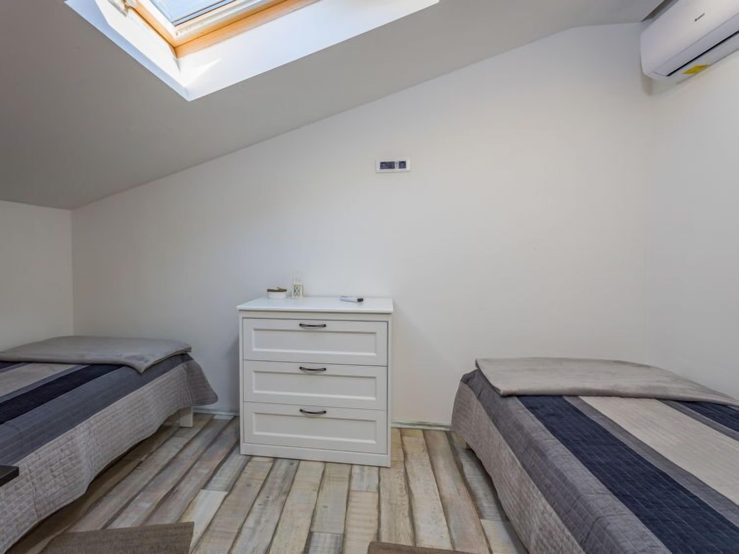 A small, modern bedroom with a skylight, a white dresser, two beds, and a wall-mounted air conditioner.