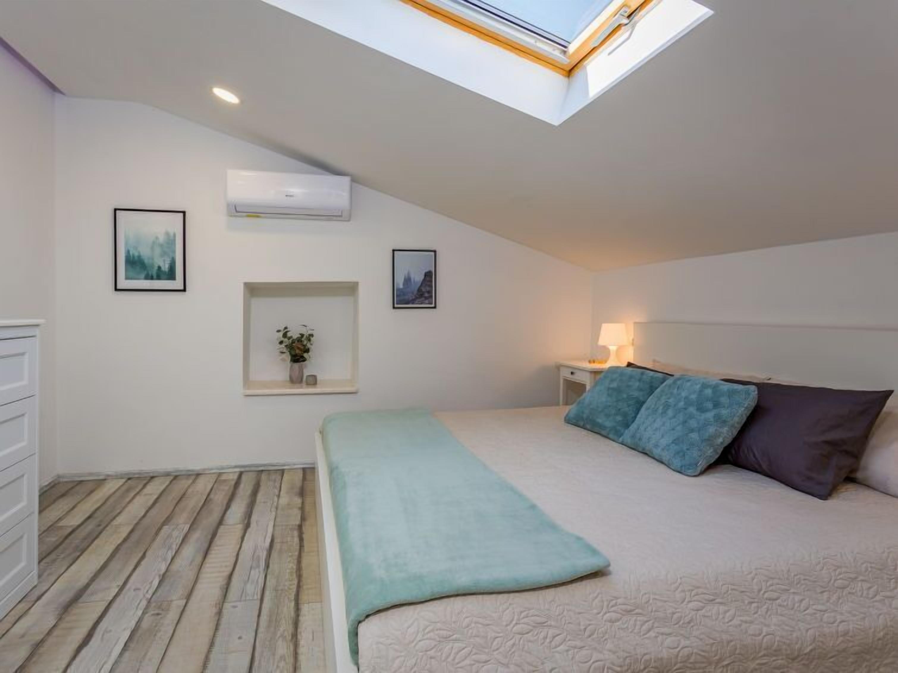 A bedroom with a skylight, a bed with a green blanket, two pillows, a bedside table with a lamp, a small shelf with a plant, and a framed picture on the wall.