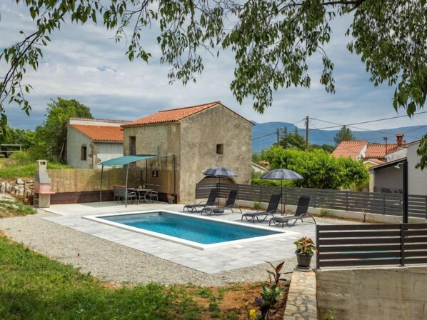 A swimming pool with lounge chairs and umbrellas, surrounded by a stone wall and a building with a red-tiled roof.
