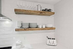 Een keuken met een witte betegelde spatwand, houten planken met borden, kopjes en een waterkoker, en een witte toaster op het aanrecht.