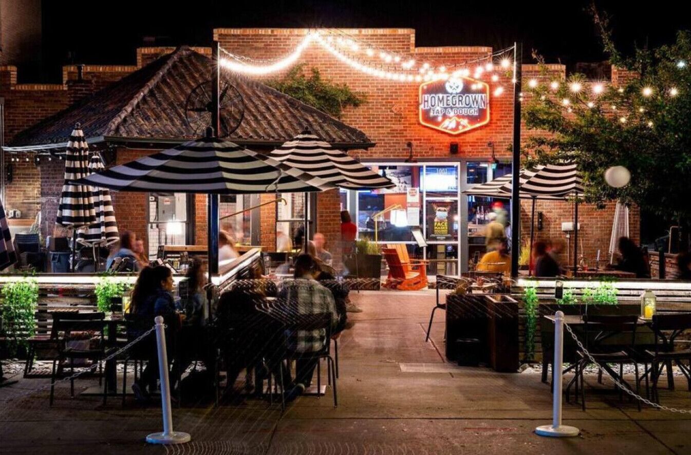 An outdoor dining area with tables and chairs, a brick building with a neon sign, and string lights.
