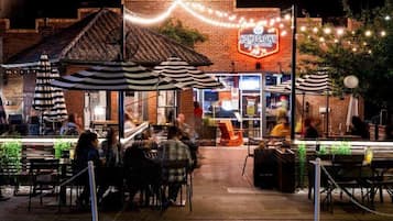 An outdoor dining area with tables and chairs, a brick building with a neon sign, and string lights.
