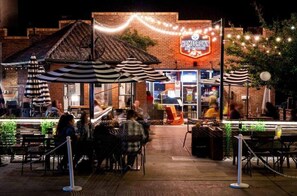 Un espace extérieur aménagé pour les repas, avec des tables et des chaises, un bâtiment en briques orné d'une enseigne lumineuse au néon et des guirlandes lumineuses.