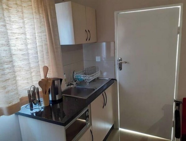 A compact kitchen with white cabinets, a stainless steel sink, and a countertop with a kettle and utensils.