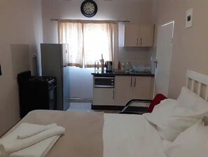 A hotel room with a bed, a small kitchenette, a refrigerator, and a clock on the wall.