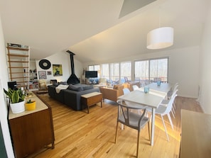A modern living room with a fireplace, a dining area, and large windows. - Private hot tub, wooded, EV charger, 1-gig fiber internet (Berkeley Springs)