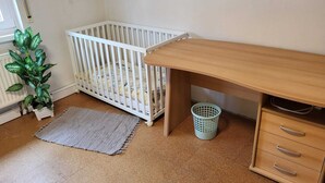 A room with a crib, a wooden desk, a small plant, a grey rug, and a laundry basket. - Large bright vacation apartment_only for private guests_ no fitters (St. Leon-Rot)
