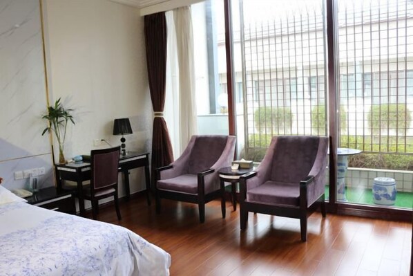 A hotel room with a bed, two armchairs, a desk, a lamp, and a view of the outside through large windows.
