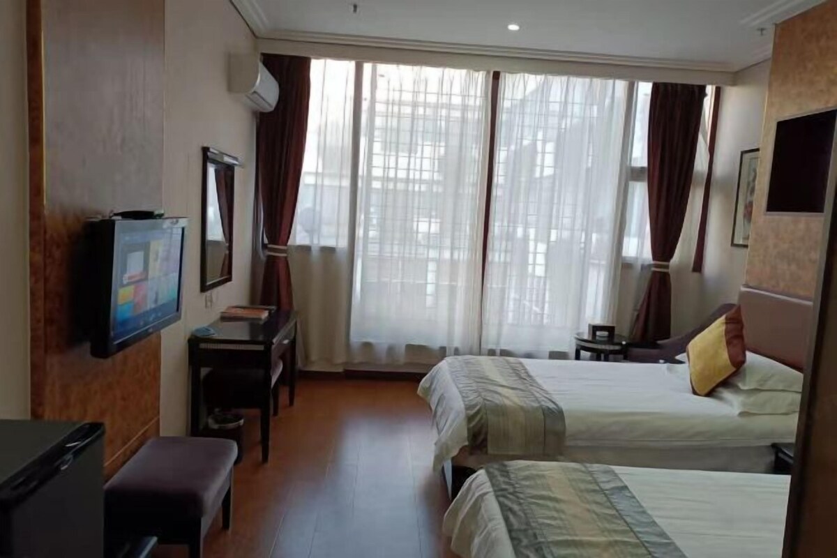 Quarto de hotel com duas camas, uma escrivaninha com cadeira, televisão e janelas amplas com cortinas.