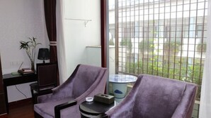 A room with two purple armchairs, a small round table, a lamp, and a view of a garden through a window with a metal grid.