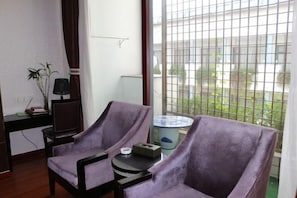 A room with two purple armchairs, a small round table, a lamp, and a view of a garden through a window with a metal grid. - Rongma Hotel (Huai'an)