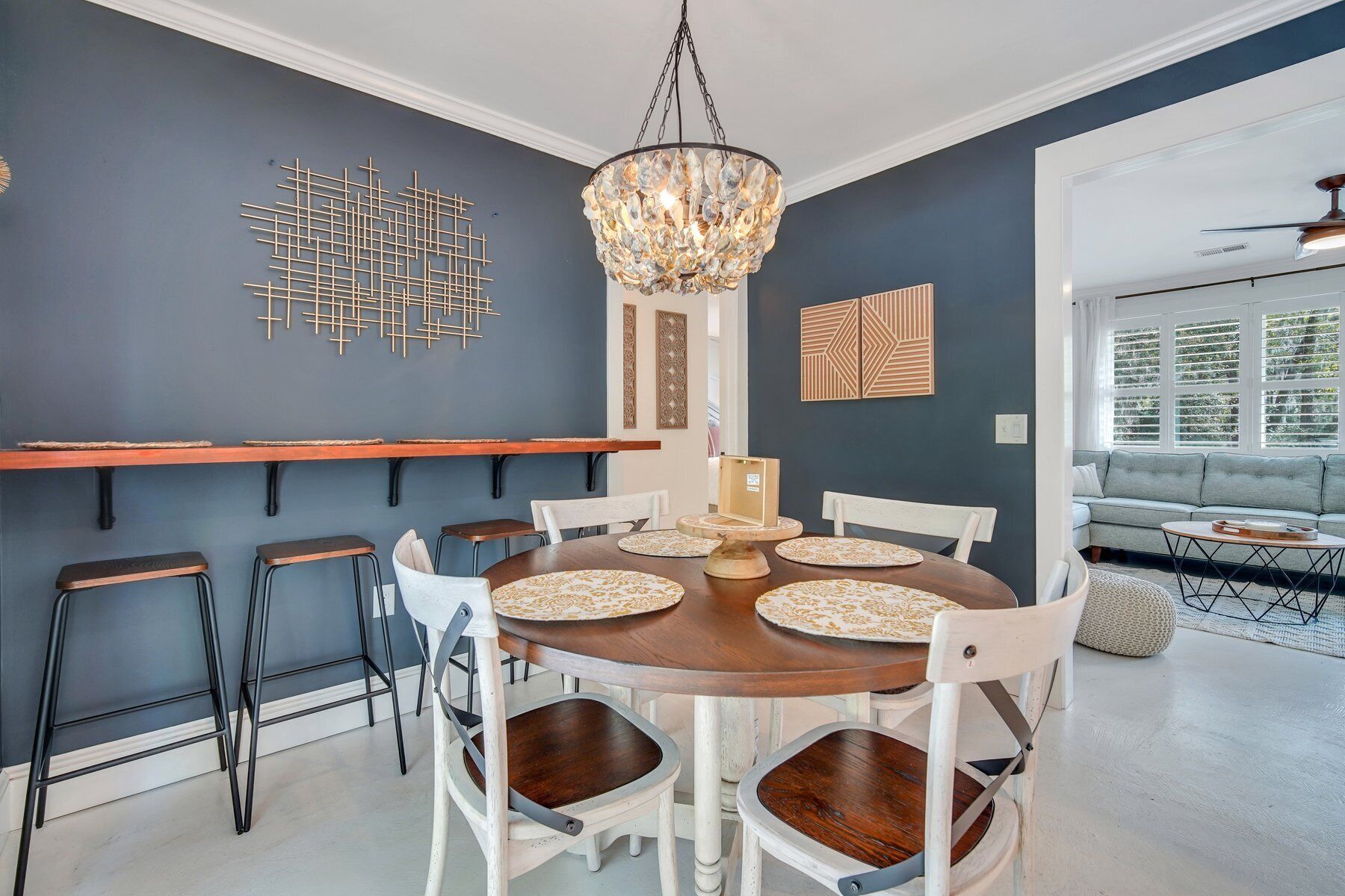 Un comedor con una mesa redonda de madera, sillas blancas y una araña. Se observan elementos decorativos en la pared y una ventana con persianas.