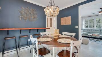 Un comedor con una mesa redonda de madera, sillas blancas y una araña. Se observan elementos decorativos en la pared y una ventana con persianas.