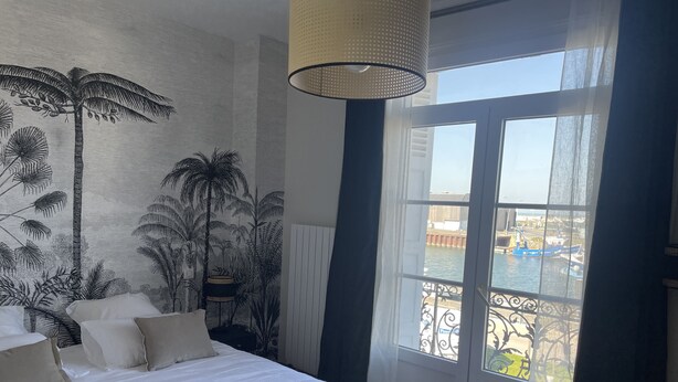 A bedroom with a large bed, a mural of tropical plants, a window with a view of boats, and a hanging light fixture. - Villa quai du Treport 4* facing port and sea. (Le treport)