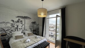 A bedroom with a large bed, a bench, a desk, and a view of the city through a large window. - Villa quai du Treport 4* facing port and sea. (Le treport)