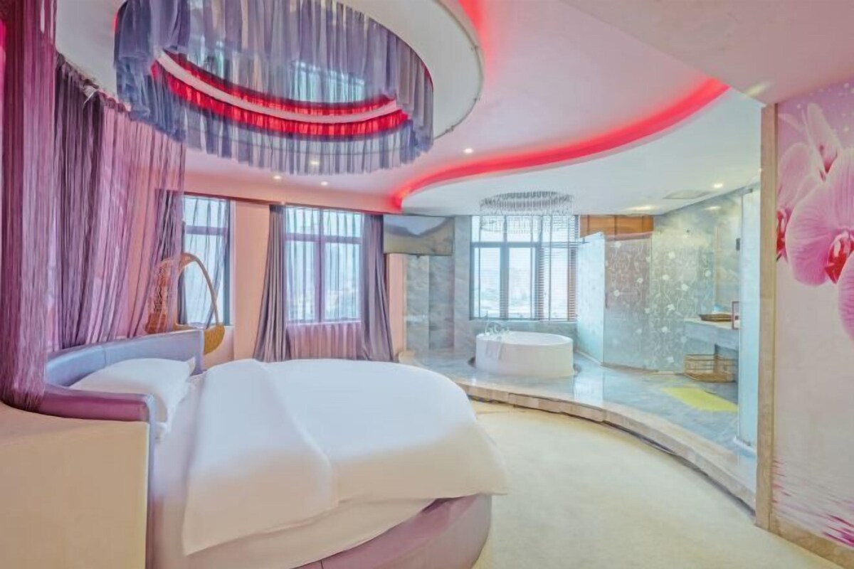 A modern bedroom with a curved bed, a large window, and a bathroom with a bathtub.