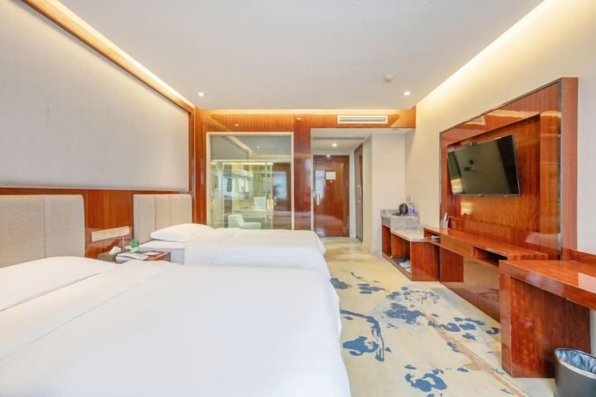 A hotel room with two beds, a TV, a desk, and a bathroom visible through a glass door.