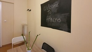 Une pièce avec un tableau noir sur lequel on peut lire « Bienvenue à Athènes », une chaise blanche, une chaise noire et une plante en pot.