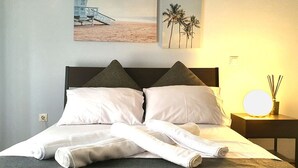 Un lit bien fait, avec des oreillers, une table de chevet avec une lampe et un mur orné d’un tableau représentant une plage et des palmiers.