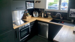 A modern kitchen with dark cabinetry, a wooden countertop, a built-in oven, a dishwasher, and a window with a view of parked cars outside. - T4 family home 3 km from the beach (Anglet)