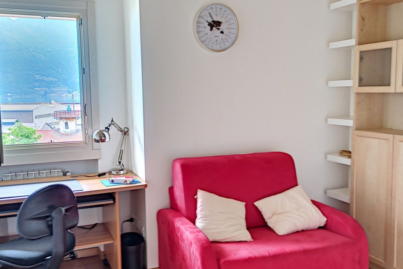 Ein Zimmer mit Schreibtisch, einem roten Sessel, einer Wanduhr und einem Fenster mit Blick auf Gebäude und Berge.