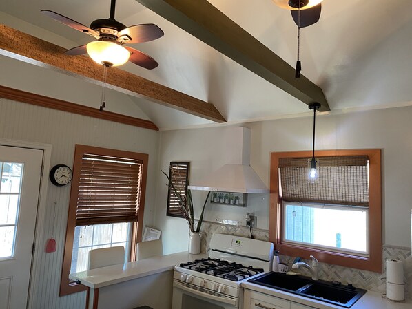 A kitchen with a gas stove, oven, and a ceiling fan. There are windows with blinds, a clock on the wall, and a small plant on the counter. - Contemporary Cottage in the heart of Mandeville (Mandeville)