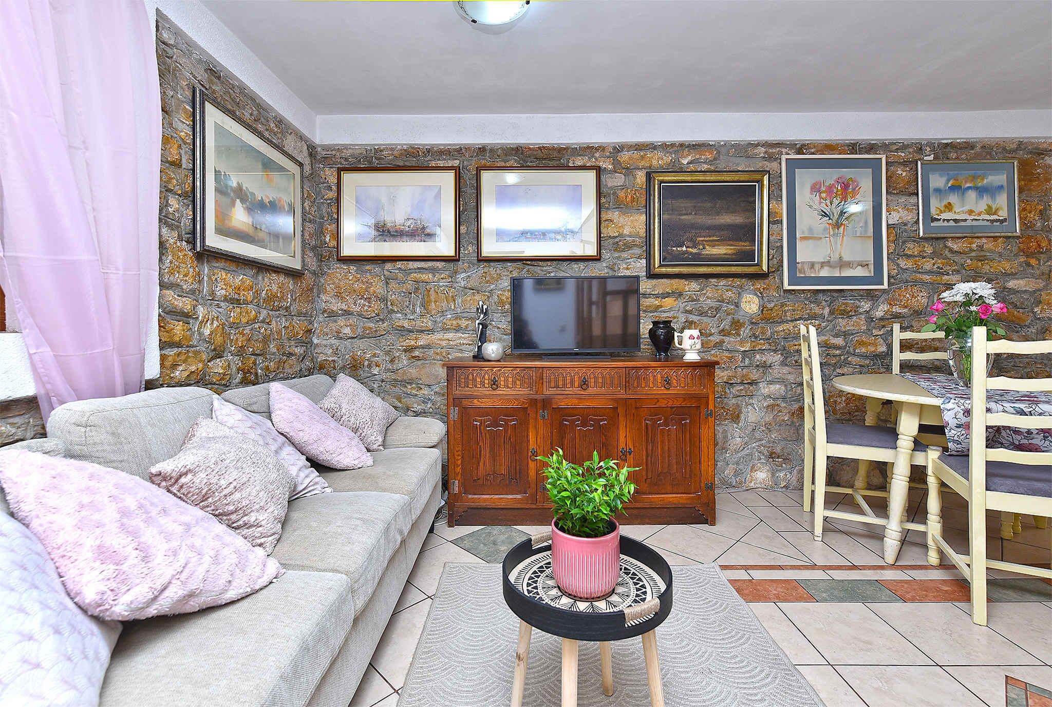 A living room with a sofa, a coffee table with a plant, a TV, and a cabinet against a stone wall with framed pictures.