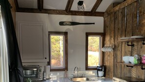 A modern kitchen with white cabinets, a stainless steel refrigerator, a microwave, and a coffee maker. There are two windows providing natural light, and a wooden ceiling with exposed beams.