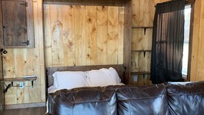 A wooden cabin interior with a bed, leather sofa, and a wooden ceiling with exposed beams.