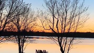 A sunset view with two chairs facing a calm lake, surrounded by leafless trees.