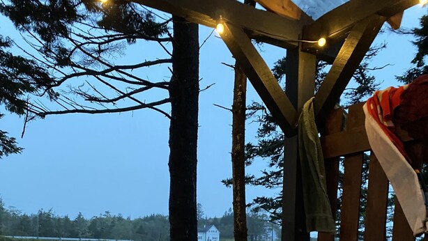 A hot tub with a wooden structure overhead, surrounded by trees and a view of a house in the background.
