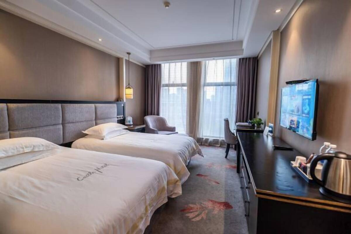 Ein Hotelzimmer mit zwei Betten, einem Schreibtisch, einem Stuhl, einem Fernseher und einem großen Fenster mit Vorhängen.
