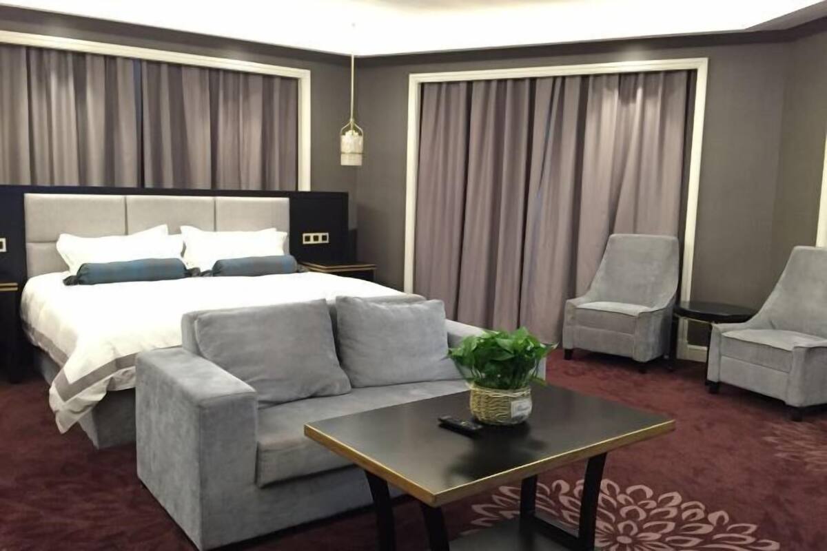 Ein Hotelzimmer mit Bett, grauem Sofa, Sesseln, einem kleinen Tisch und einer Pflanze.
