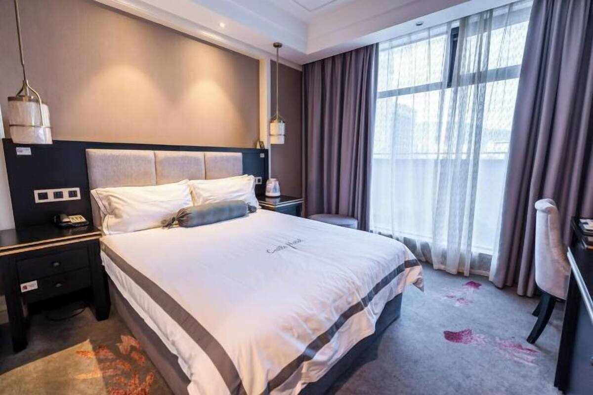 Ein Hotelzimmer mit einem großen Bett, einem Schreibtisch und einem Stuhl. Es gibt ein Fenster mit Vorhängen und Blick auf die Stadt.