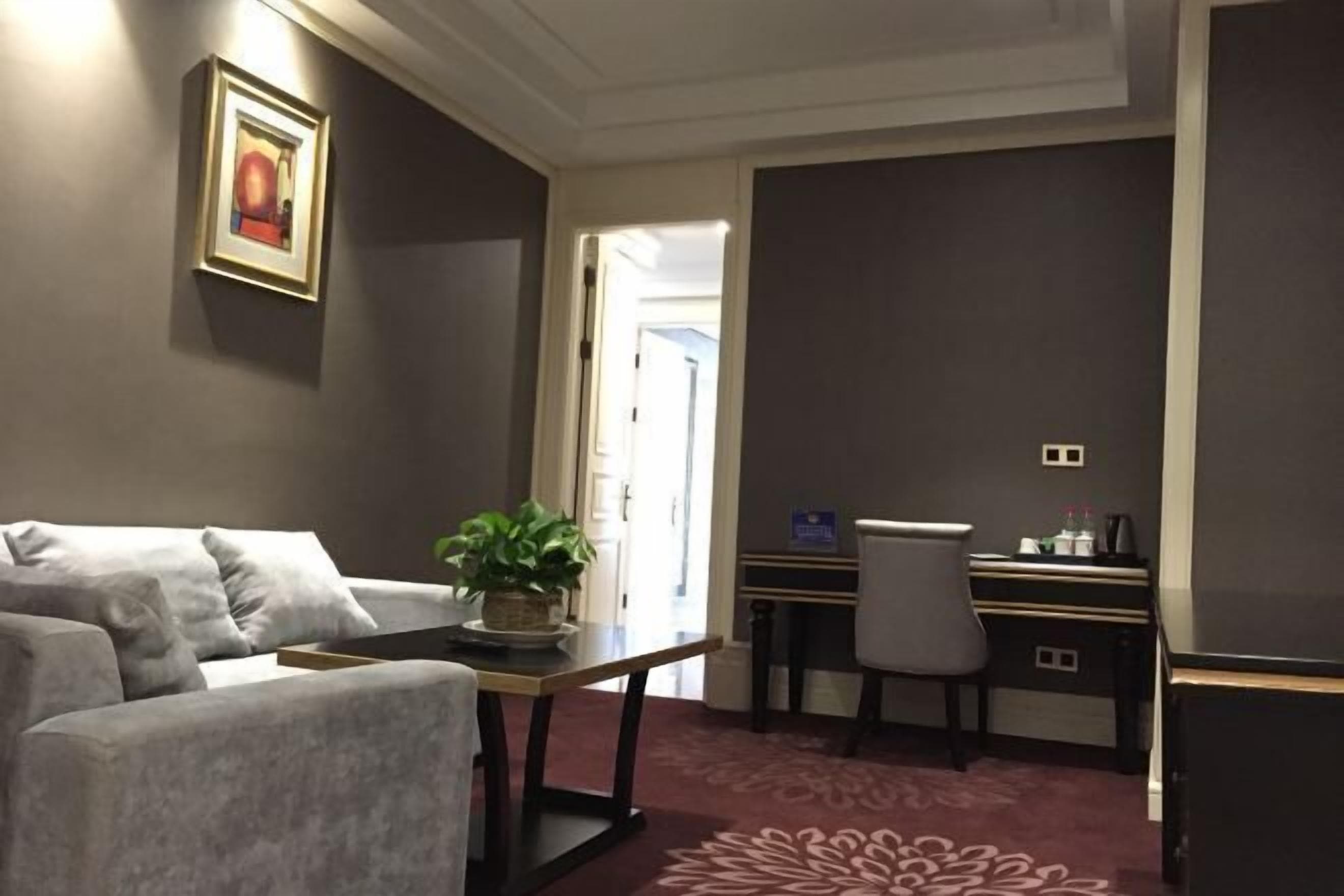 A modern hotel room with a grey sofa, a small table with a plant, a desk with a chair, and a carpeted floor.