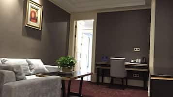 A modern hotel room with a grey sofa, a small table with a plant, a desk with a chair, and a carpeted floor.