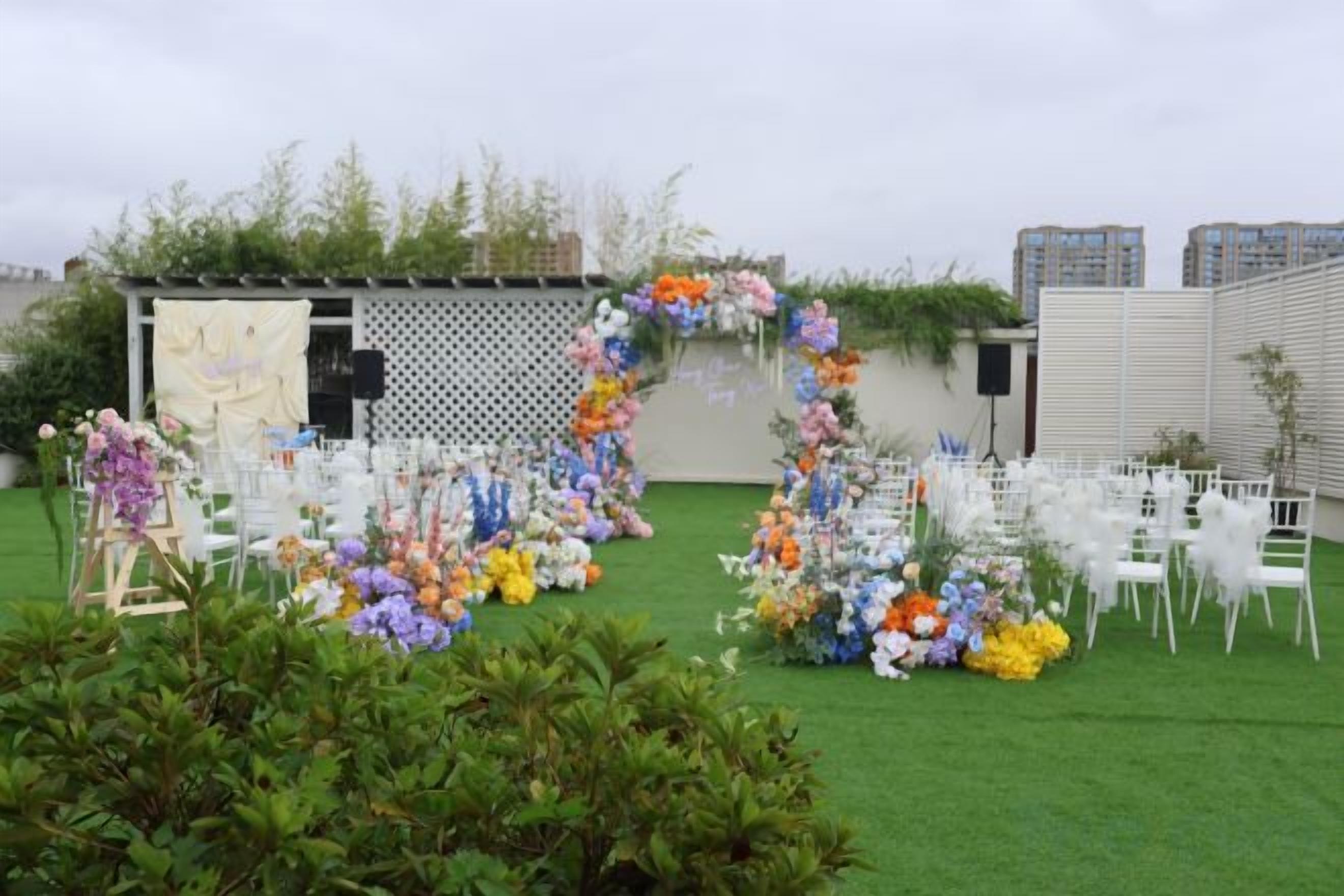 An outdoor event space with chairs arranged in rows, a floral archway, and a backdrop with a white fence and greenery.