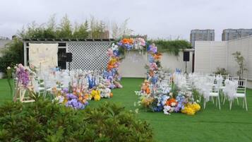 An outdoor event space with chairs arranged in rows, a floral archway, and a backdrop with a white fence and greenery.