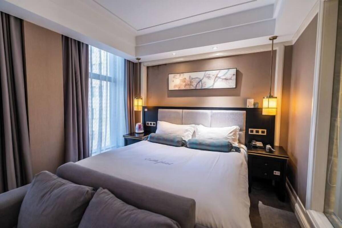 Ein modernes Hotelzimmer mit einem großen Bett, zwei Nachttischen mit Lampen, einer Kopfstütze mit einem Gemälde und Blick ins Freie durch große Fenster.