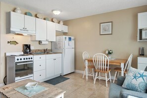A compact kitchen with white cabinets, a stove, and a refrigerator. There is a dining area with a wooden table and white chairs.