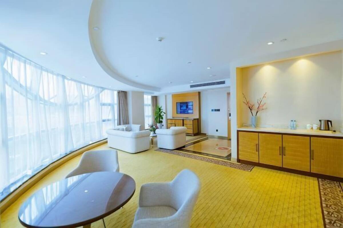 A modern hotel room with a large window, a dining area with a table and chairs, a sofa, a kitchen area with cabinets and a TV, and a yellow patterned carpet.