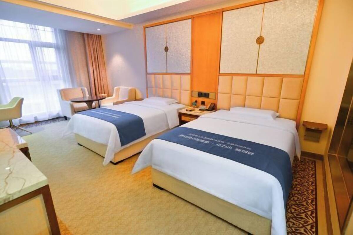 A hotel room with two beds, a desk, a chair, and a nightstand with a lamp.