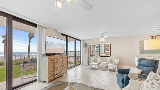 A spacious living room with a large TV, comfortable seating, a ceiling fan, and a view of the beach through large windows. - Direct Oceanfront, Ground-Floor Condo Just Steps from Pool & Beach Access! (New Smyrna Beach)