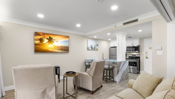 A modern living room with a sofa, armchairs, a coffee table, and a TV. There is a kitchen area with a microwave and oven in the background. - MUIRFIELD 2820 AT INNISBROOK AVAILABLE FOR THE 2027 VALSPAR PGA TOURNAMENT (Palm Harbor)