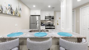 A modern kitchen with white cabinets, a stainless steel refrigerator, a microwave, and an oven. There are two blue placemats on the marble countertop. - MUIRFIELD 2820 AT INNISBROOK AVAILABLE FOR THE 2027 VALSPAR PGA TOURNAMENT (Palm Harbor)