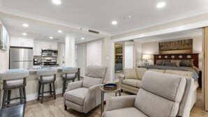 A modern living room with a sofa, armchairs, and a coffee table. There is a kitchen area with a refrigerator, microwave, and oven. The room has white walls and a wooden floor. - MUIRFIELD 2820 AT INNISBROOK AVAILABLE FOR THE 2027 VALSPAR PGA TOURNAMENT (Palm Harbor)
