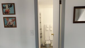 Un couloir mène à une salle de bain équipée d’un rideau de douche, d’un porte-serviettes et d’une petite table sur laquelle se trouve un panier.