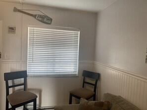 A room with a white door, a window with blinds, two dark wooden chairs, and a cushion on a light-colored sofa.