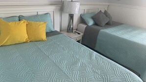 Habitación de hotel con dos camas, cada una con colcha acolchada color aguamarina y almohadas en amarillo y gris. Hay una mesita de noche con una lámpara y un control remoto.